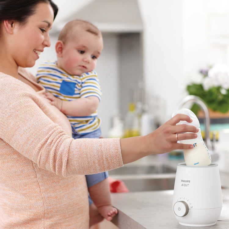Woman holding a baby and using a Philips milk warmer in a kitchen.