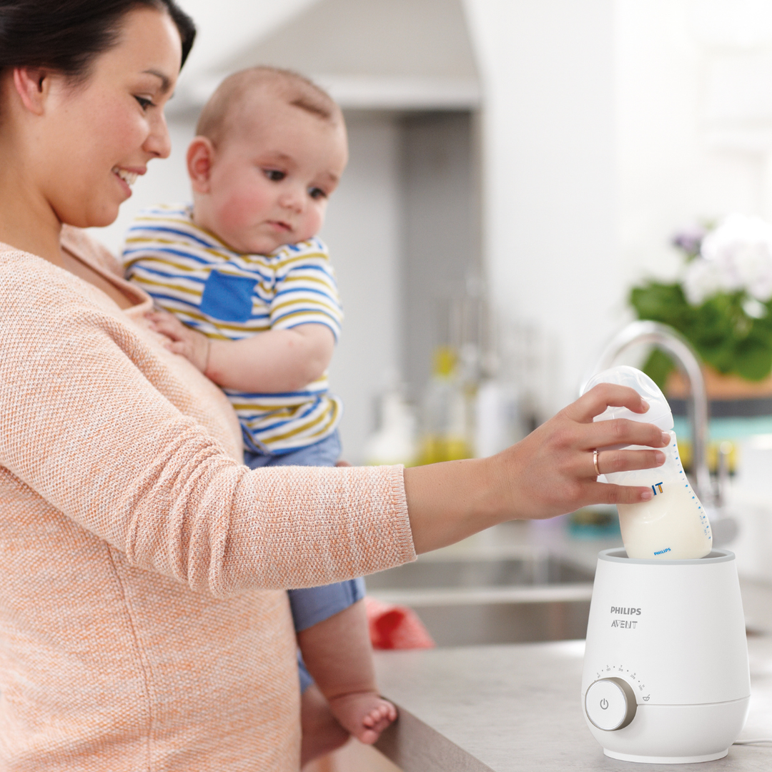 Woman holding a baby and using a Philips milk warmer in a kitchen.