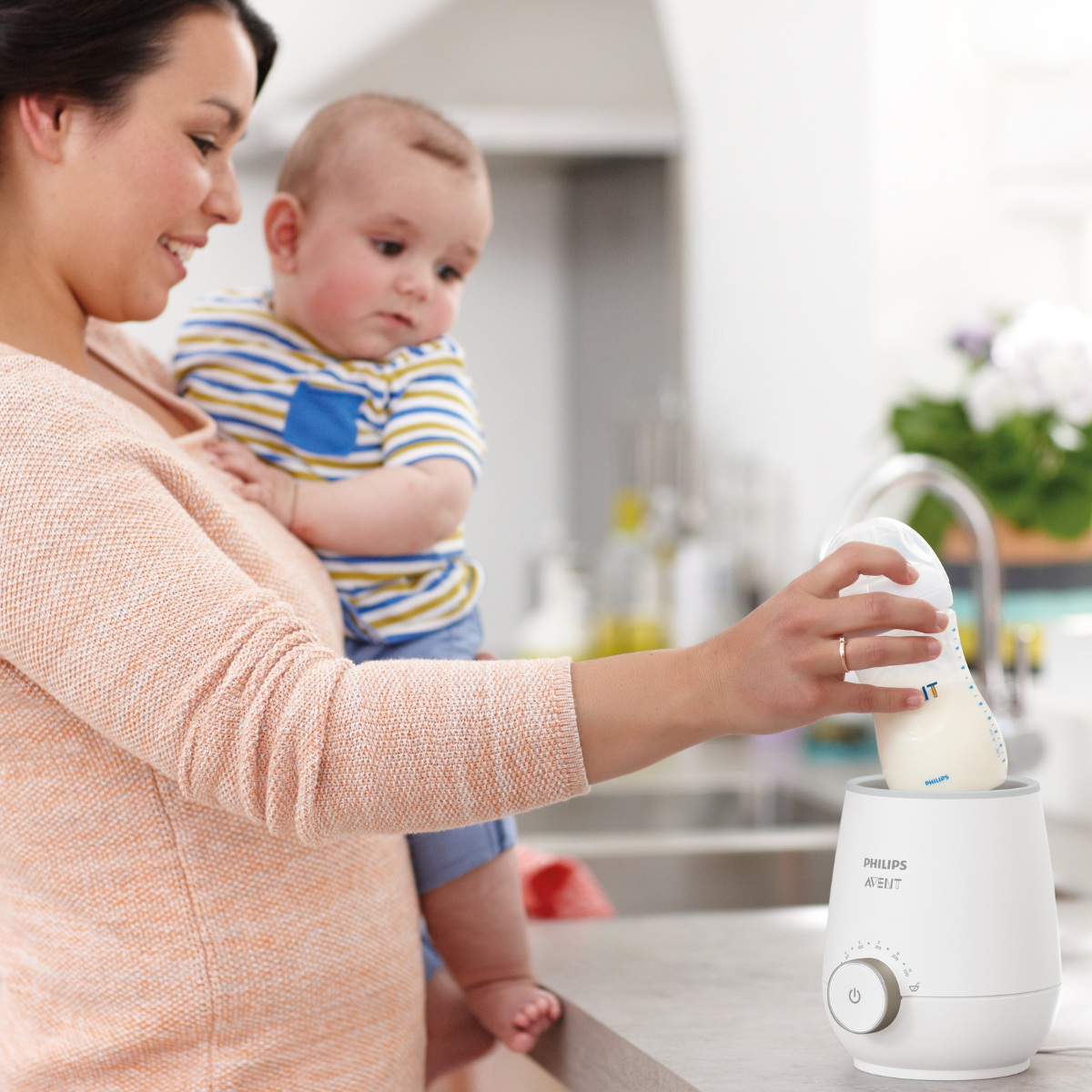 Woman holding a baby and using a Philips milk warmer in a kitchen.