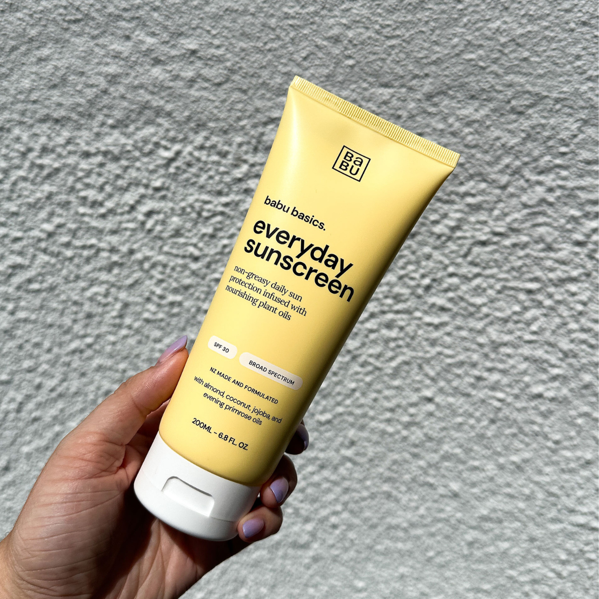 Hand holding a tube of babu basics everyday sunscreen against a gray background