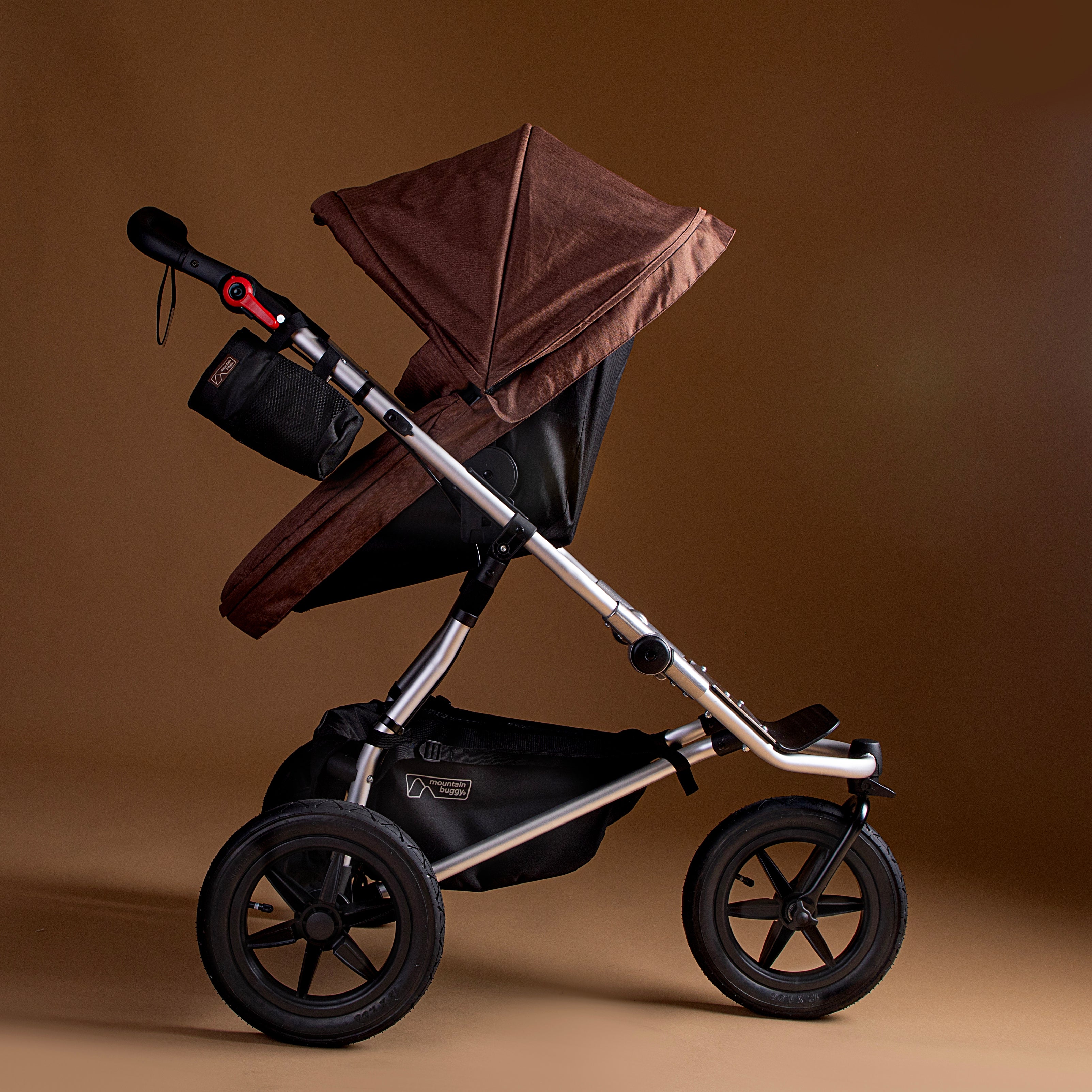 Full shot of the Mountain Buggy urban jungle carrycot plus in mustang brown in use on the urban jungle buggy shown on a brown background