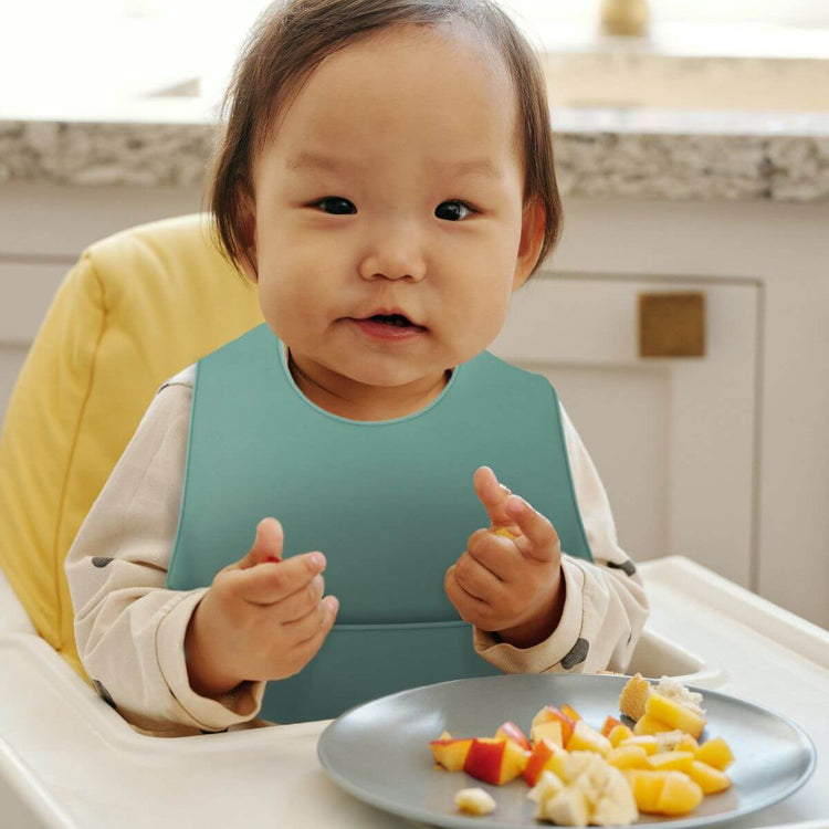 Baby wearing grey ORIGIN Silicone Bib eating at highchair with food falling into catch pocket.