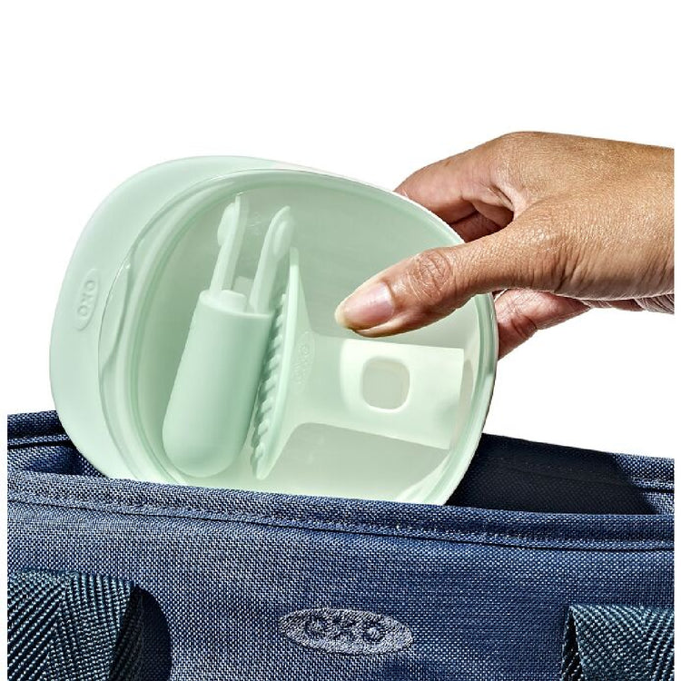 Hand opening a light green silicone container with utensils inside, held over a blue bag.