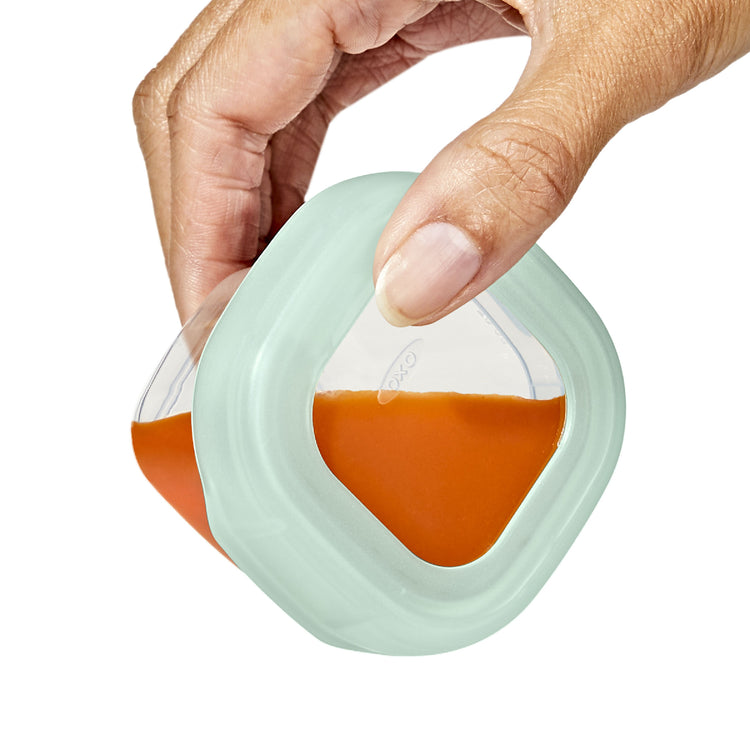Hand holding a clear container with a light green lid filled with a brown liquid on a white background
