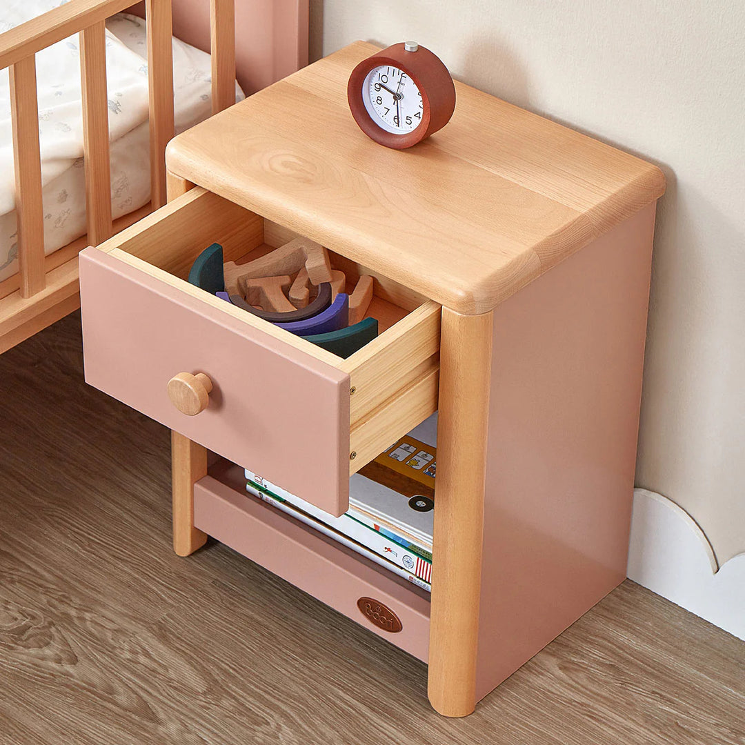 Wooden nightstand with open drawer containing toys and books, clock on top, next to a crib.