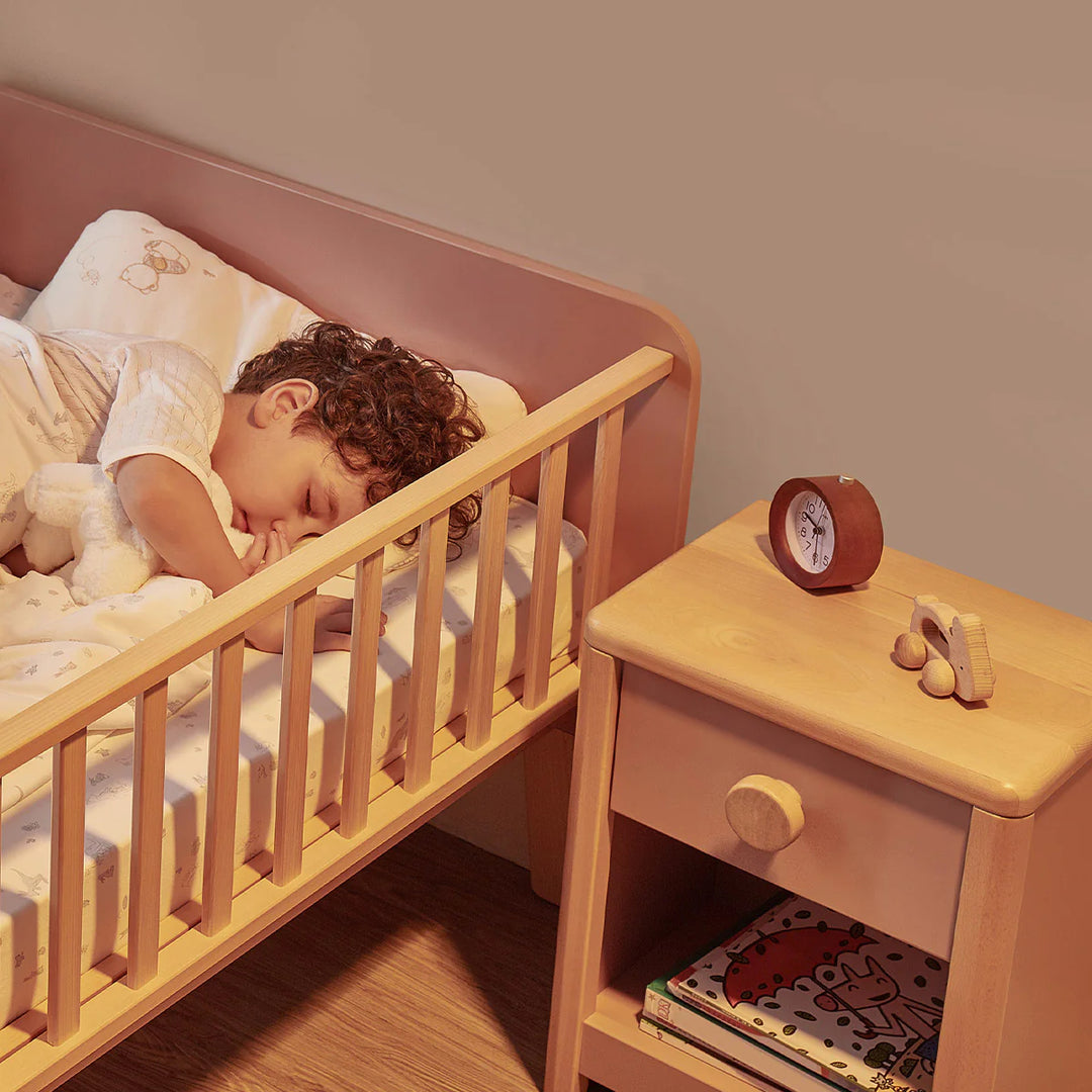 Child sleeping in a crib next to a wooden nightstand with a clock and books.