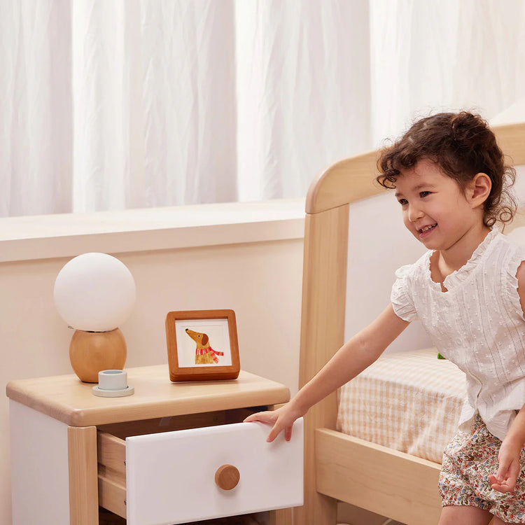 Child opening a drawer in a wooden bed with a nightstand and lamp in the background.