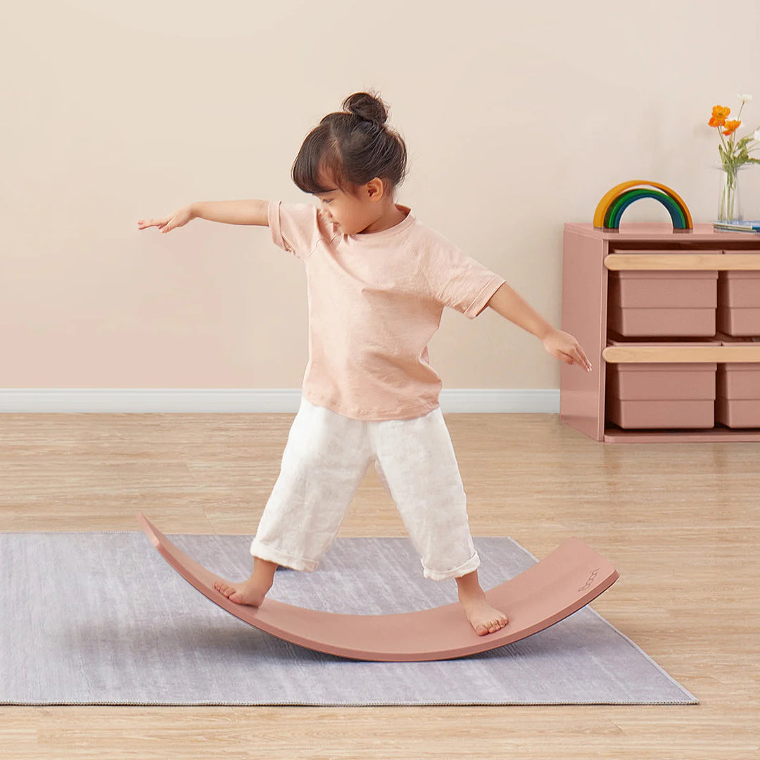 Child balancing on a pink wobble board in a room with a wooden floor and light-colored walls.