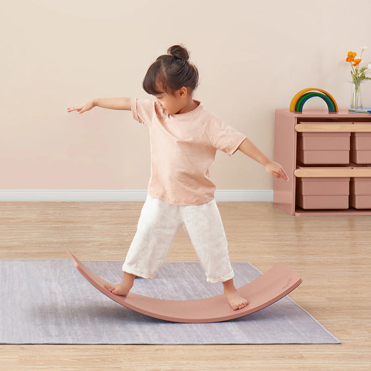 Child balancing on a pink wobble board in a room with a wooden floor and light-colored walls.
