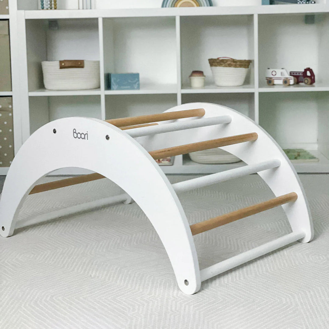 White baby arch with wooden bars on a light gray floor, with a shelf in the background.
