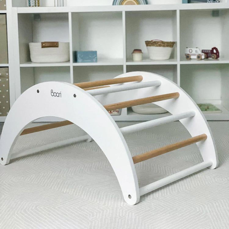 White baby arch with wooden bars on a light gray floor, with a shelf in the background.