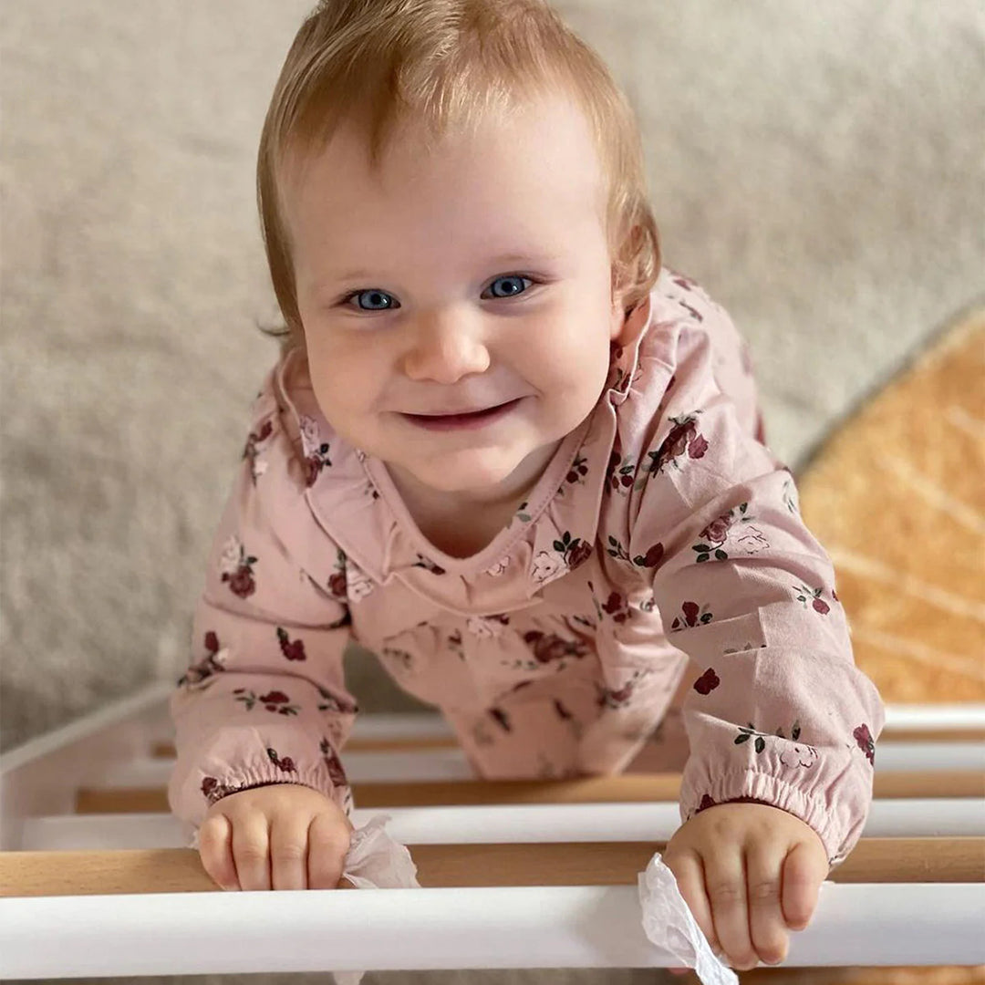 Baby in a pink floral outfit standing on a wooden floor.
