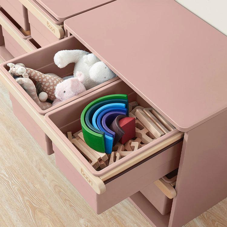 Pink toy storage unit with open drawers containing toys and wooden blocks on a light wood floor.