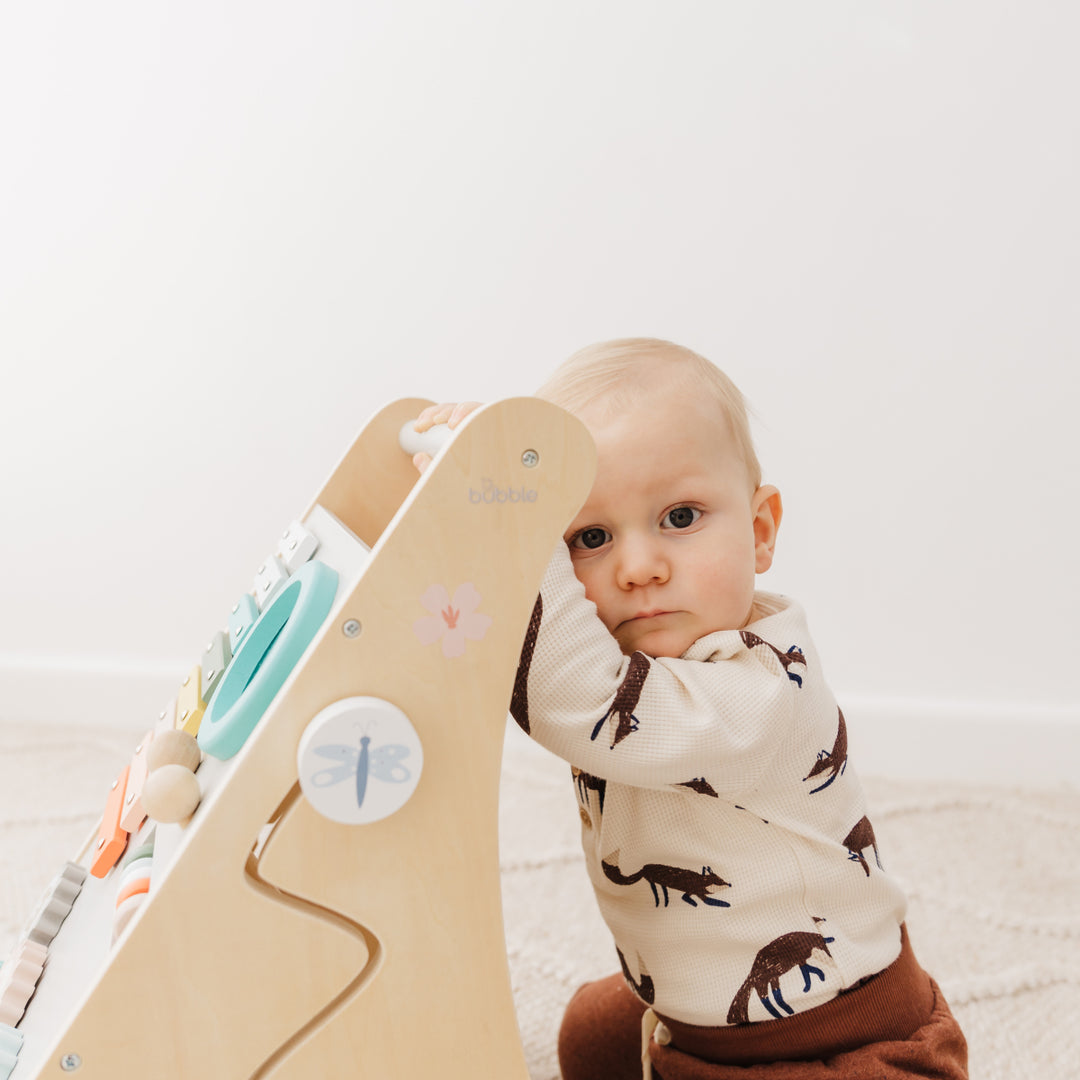 Baby in a dinosaur onesie standing next to a wooden climbing toy on a light background