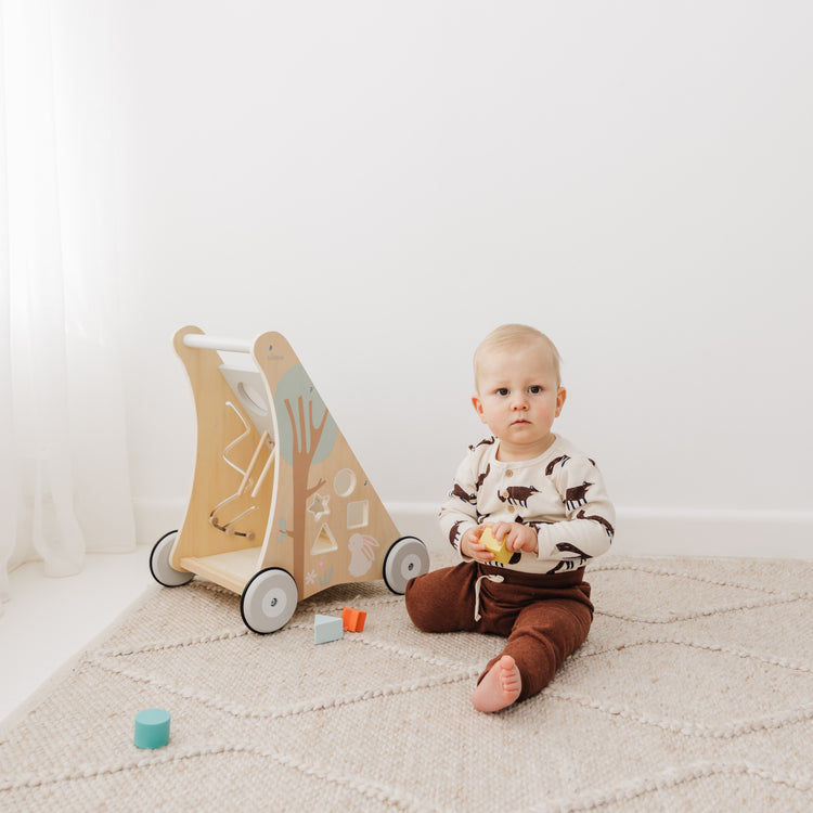 Baby sitting on a white floor with a wooden toy cart and colorful blocks.