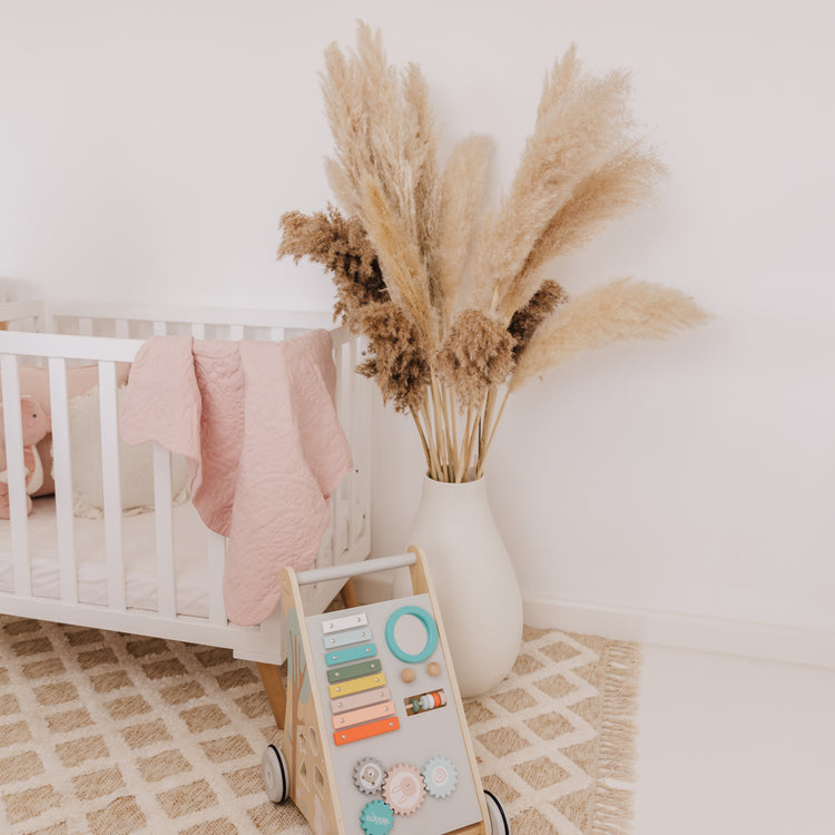 Nursery room with crib, decorative vase with pampas grass, and educational toy on a patterned rug.