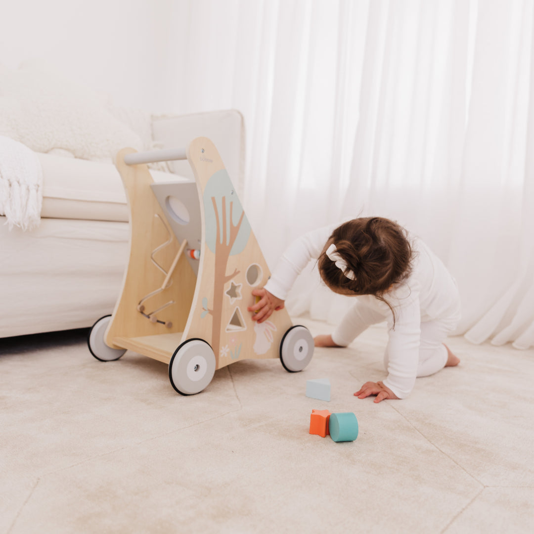 Child playing with a wooden toy walker and blocks on a light-colored floor.