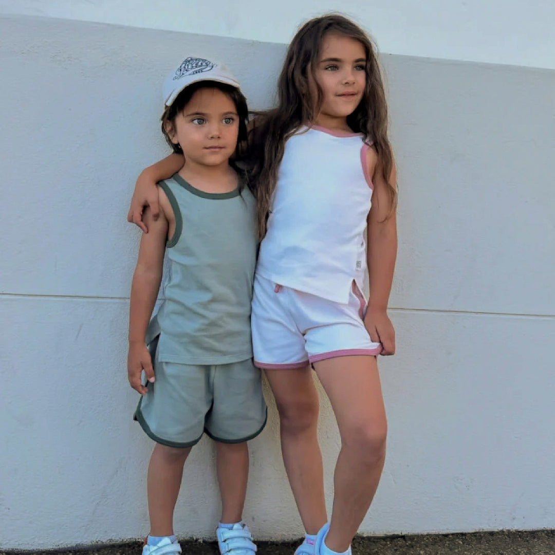 Two children standing against a white wall, one wearing a cap and the other in a white top and shorts.