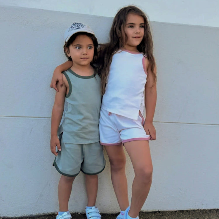 Two children standing against a white wall, one wearing a cap and the other in a white top and shorts.