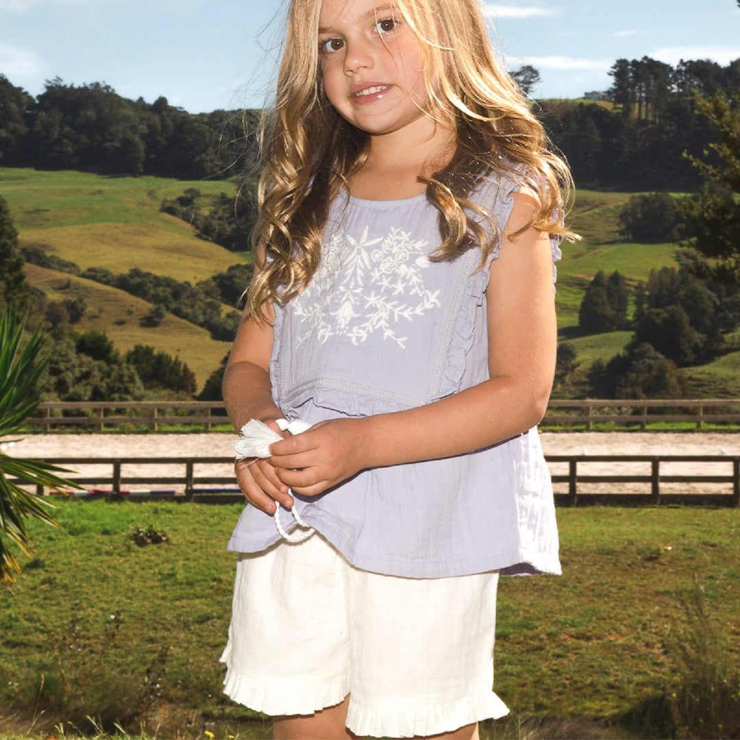 Young girl in a light blue top and white shorts standing in a grassy field with trees in the background.