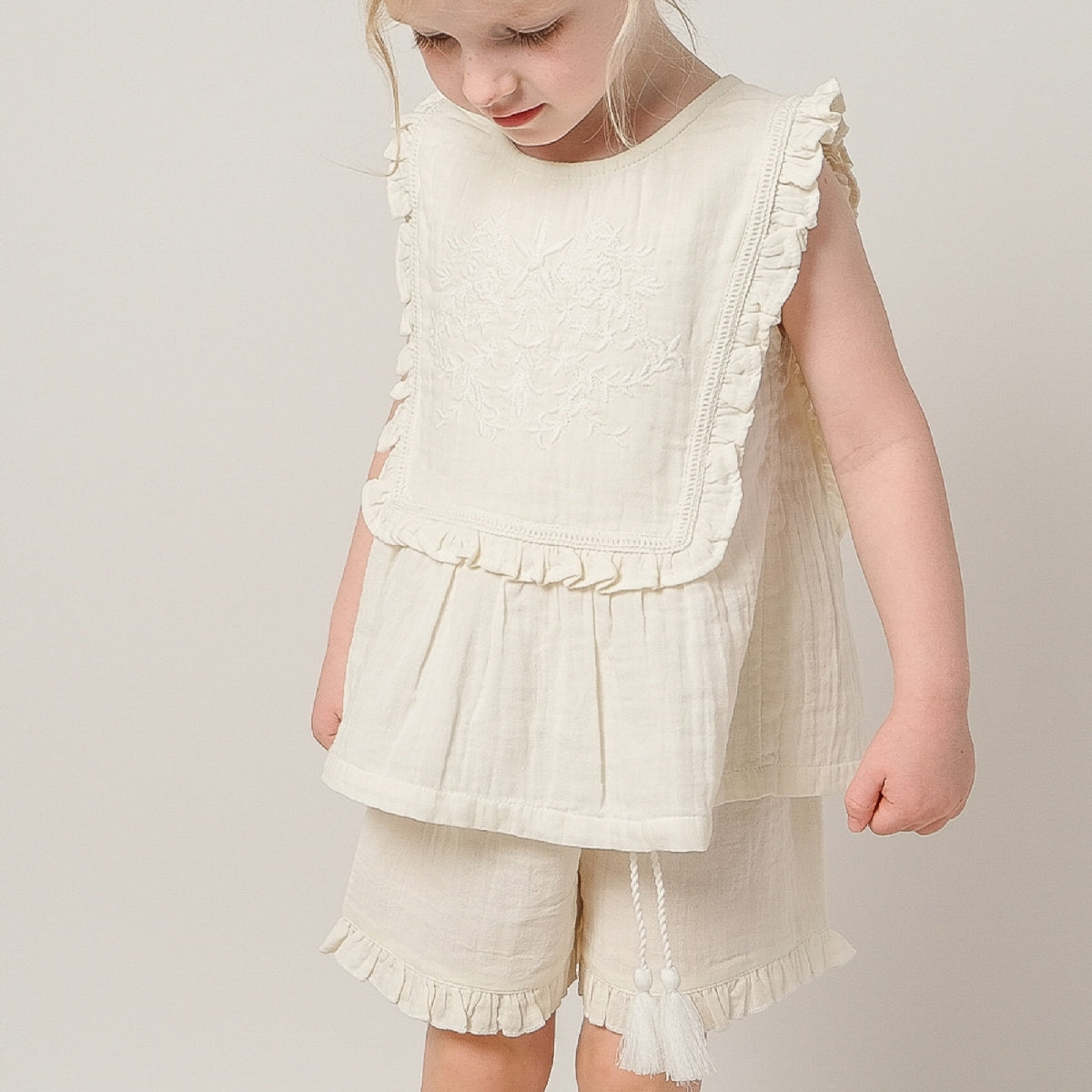 Young girl wearing a white ruffled top and shorts set on a plain background