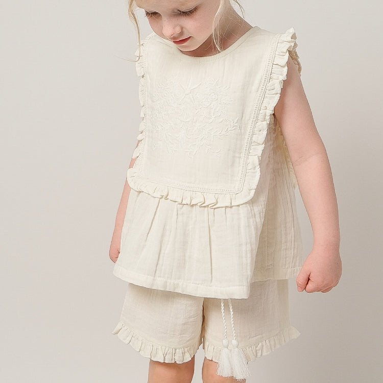 Young girl wearing a white ruffled top and shorts set on a plain background