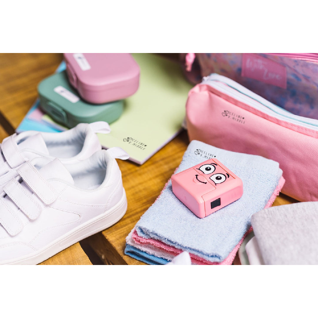 Small pink device with a cartoon face on top of folded cloths next to white sneakers and colorful bags.