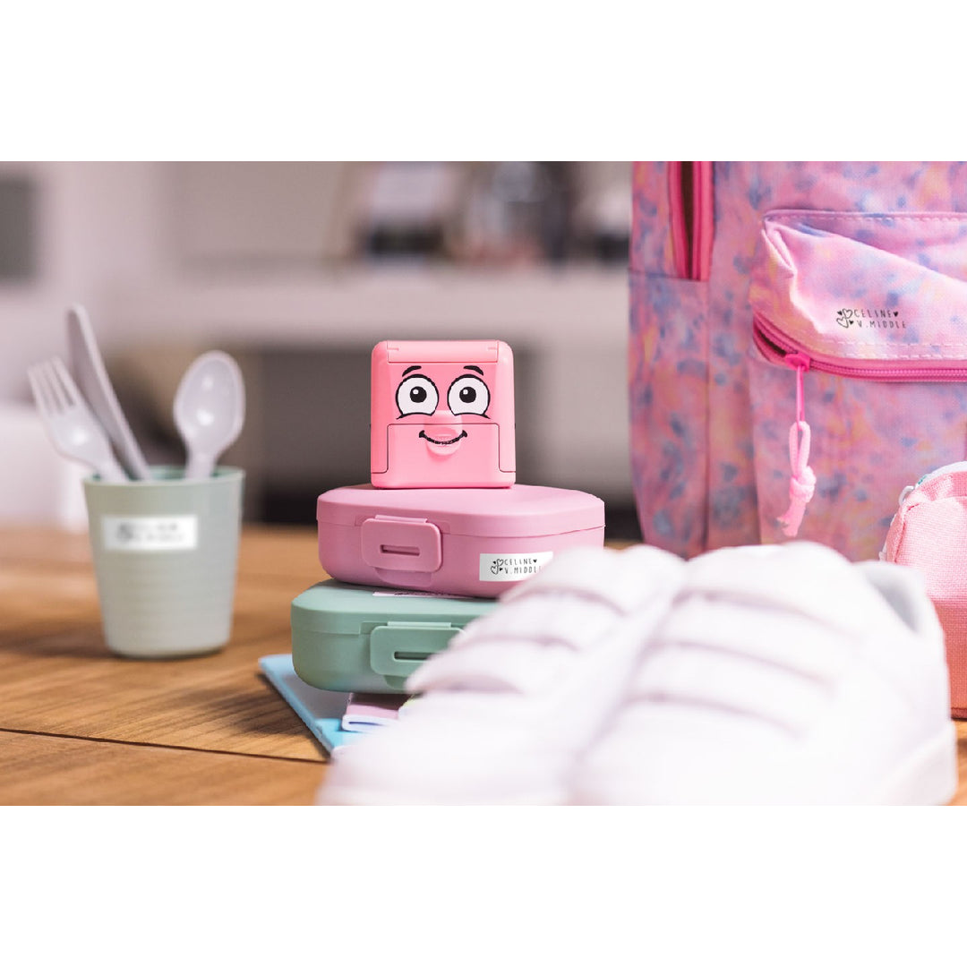 Pink lunch box with cartoon face on a table with a backpack in the background