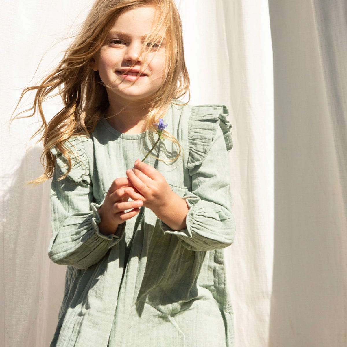 Young girl wearing a light green dress standing against a white curtain.