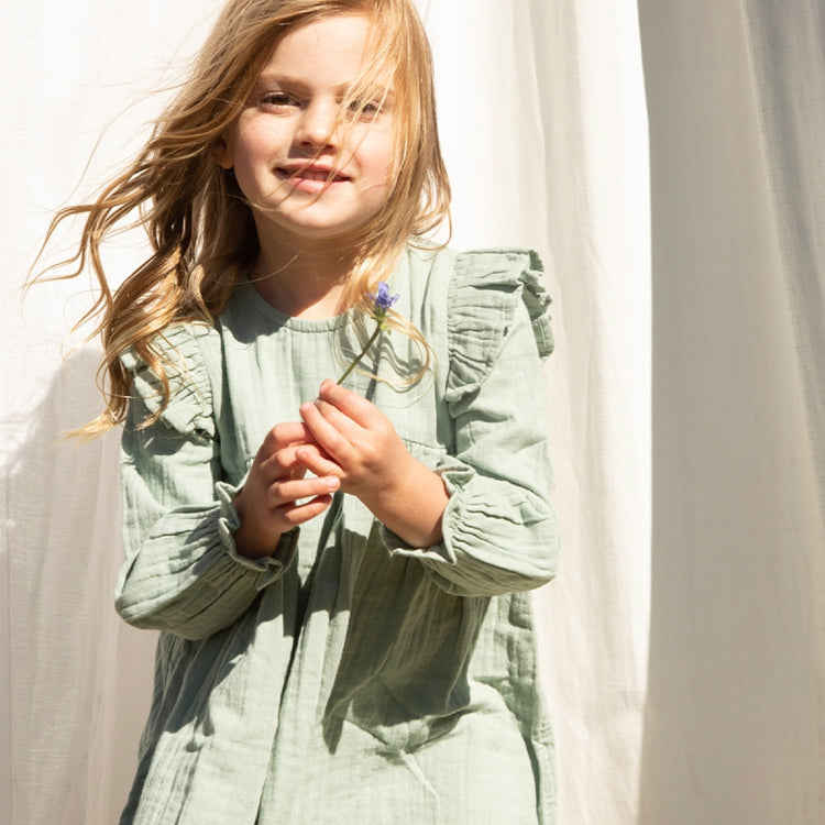 Young girl wearing a light green dress standing against a white curtain.