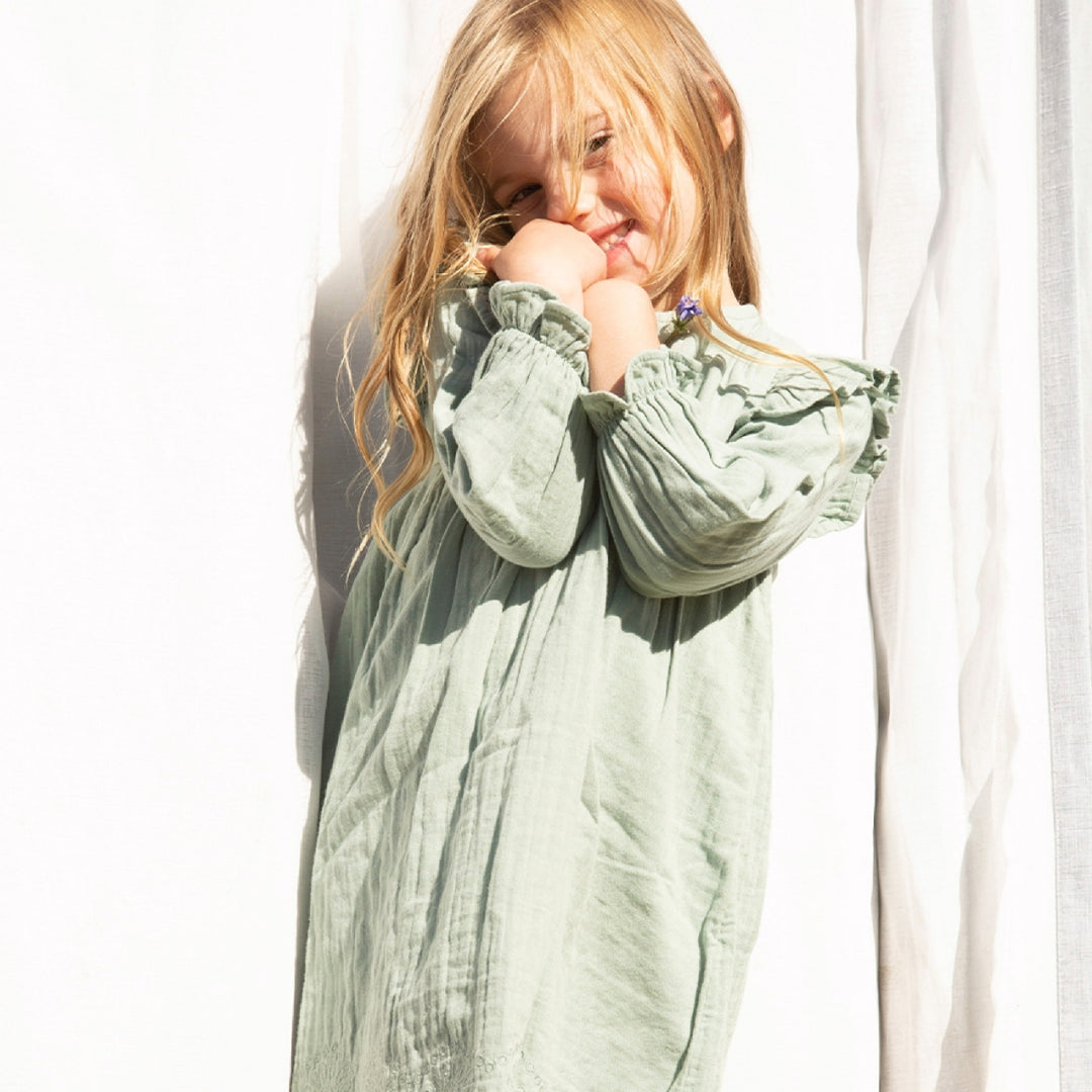 Young girl wearing a light green dress with ruffled sleeves, standing against a white curtain.