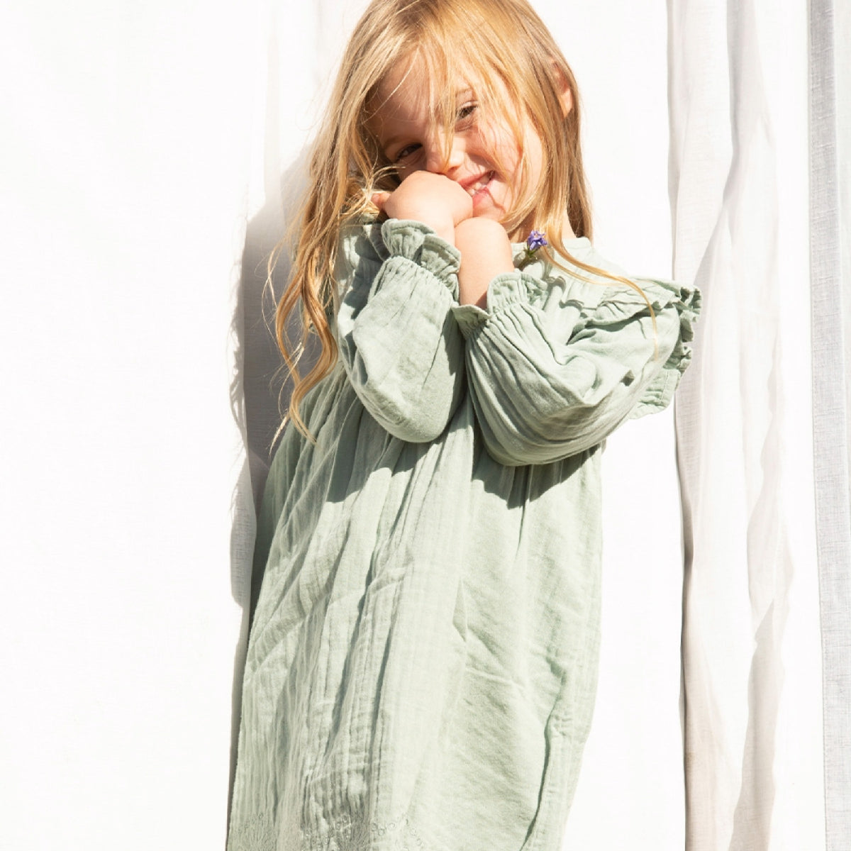 Young girl wearing a light green dress with ruffled sleeves, standing against a white curtain.