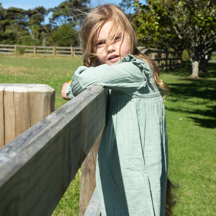 Young girl in a green dress leaning against a wooden fence in a park.
