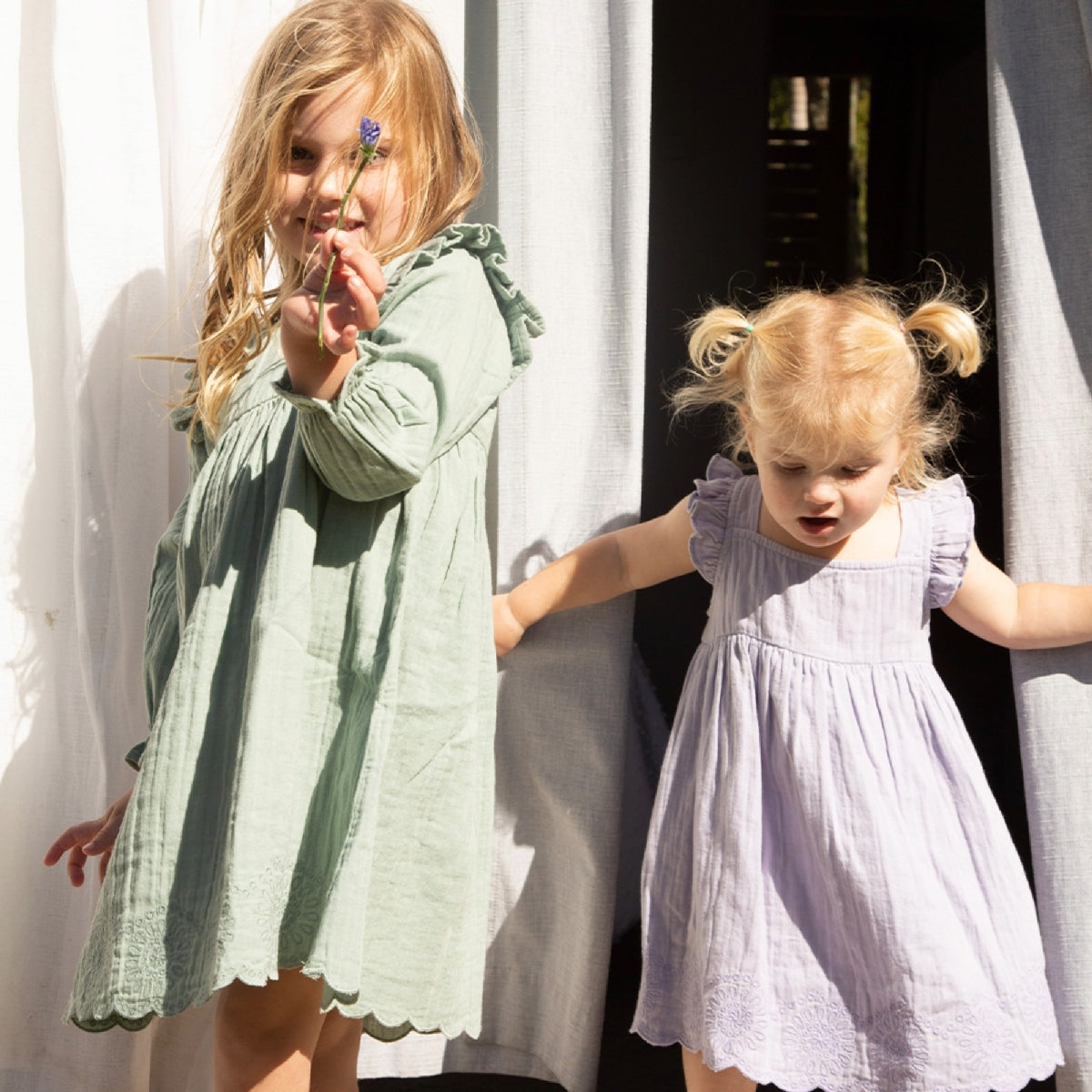 Two young girls in dresses standing outdoors with a white curtain in the background.