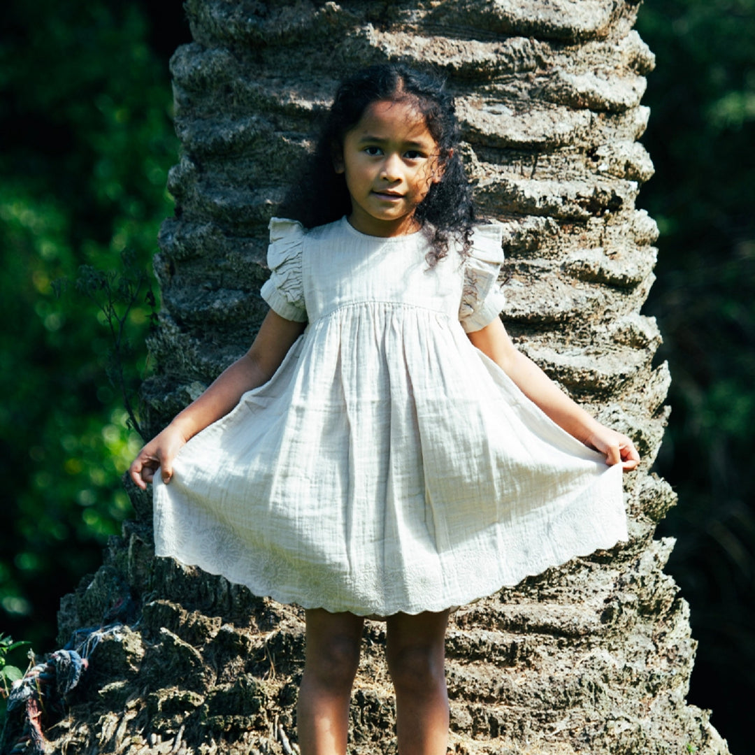Young girl in a white dress standing against a textured stone wall with greenery in the background