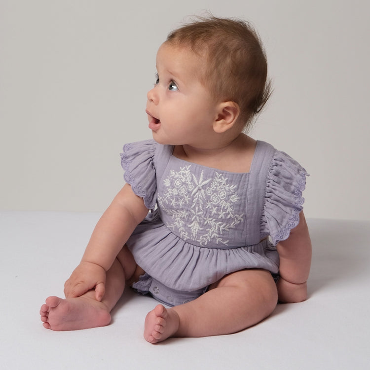 Baby wearing a purple embroidered romper sitting on a white surface.