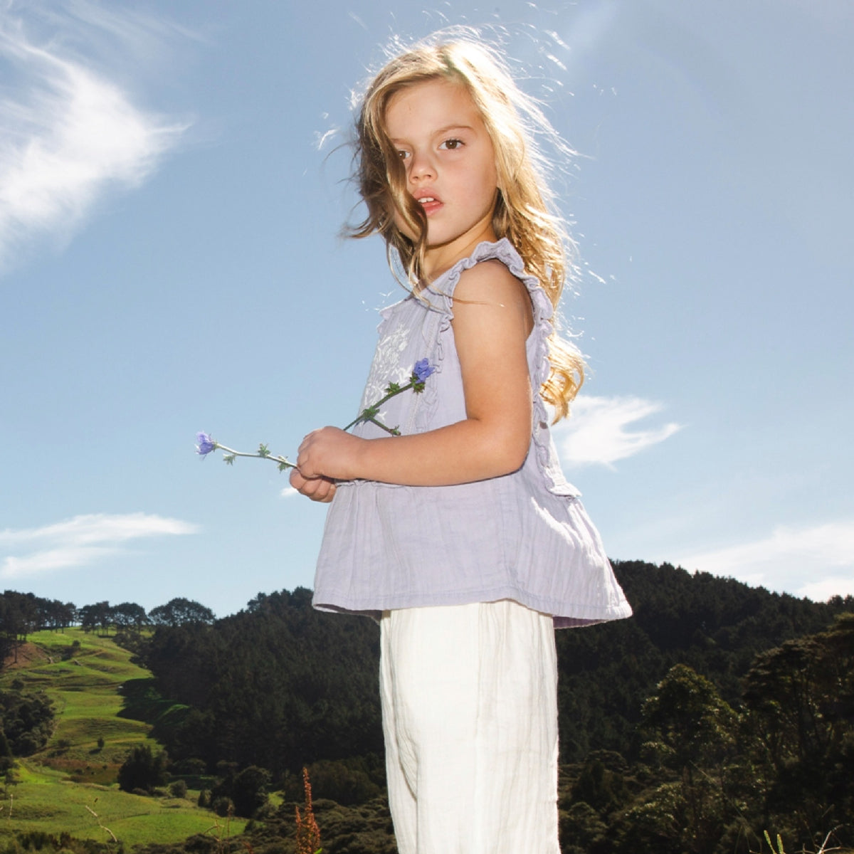 Young girl holding flowers against a scenic background with green hills and blue sky.