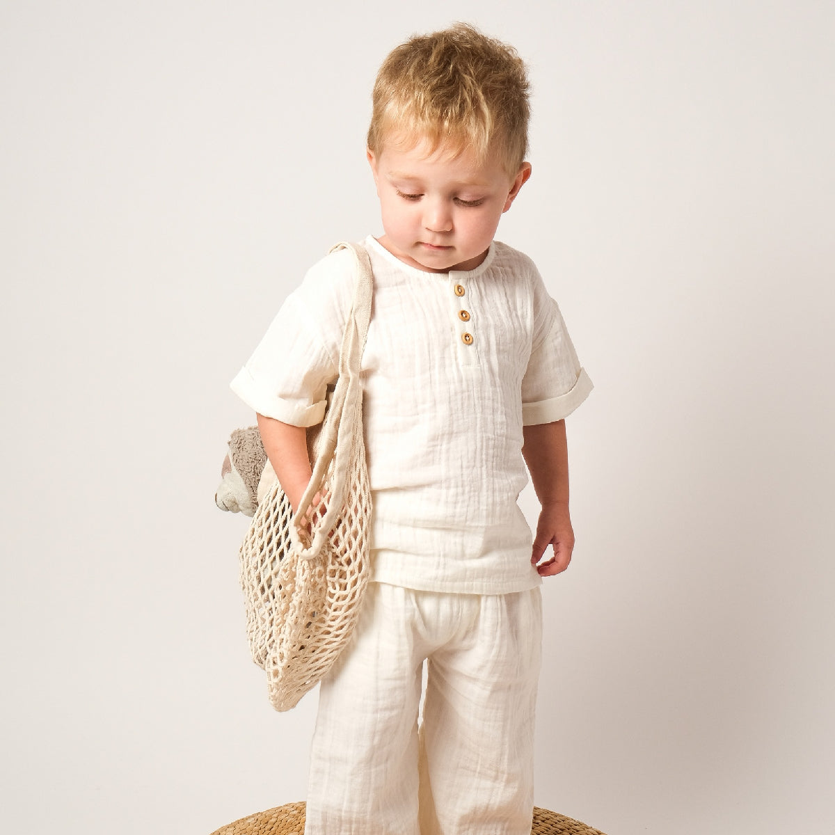 Child wearing a white outfit holding a woven bag on a plain background