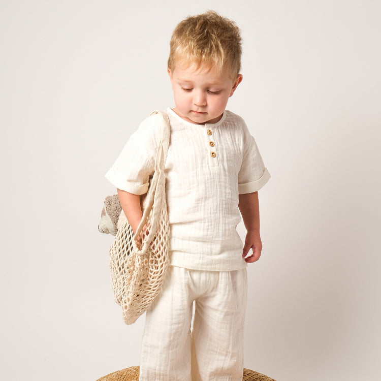 Child wearing a white outfit holding a woven bag on a plain background