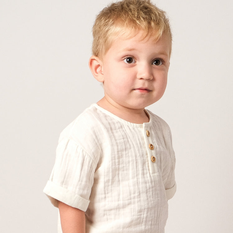 Young child wearing a white textured shirt with gold buttons on a plain background