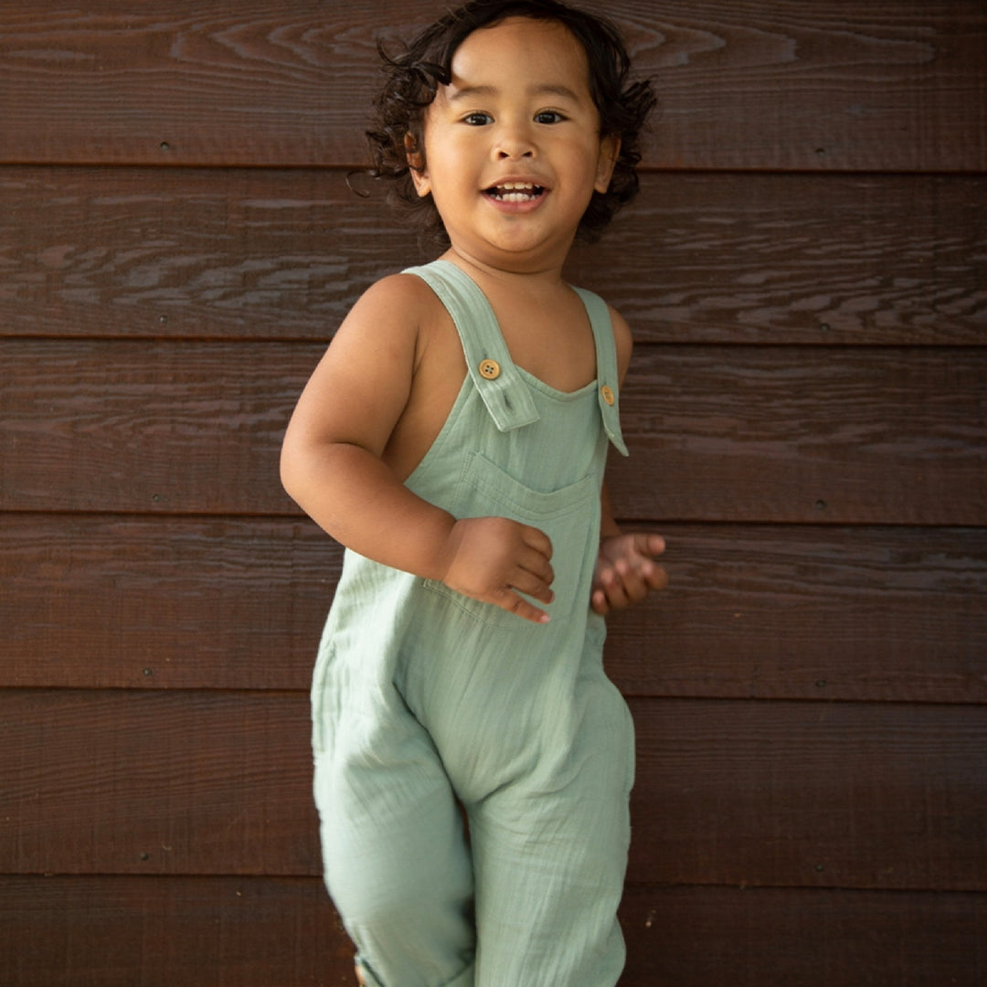 Child wearing light green overalls standing against a wooden wall