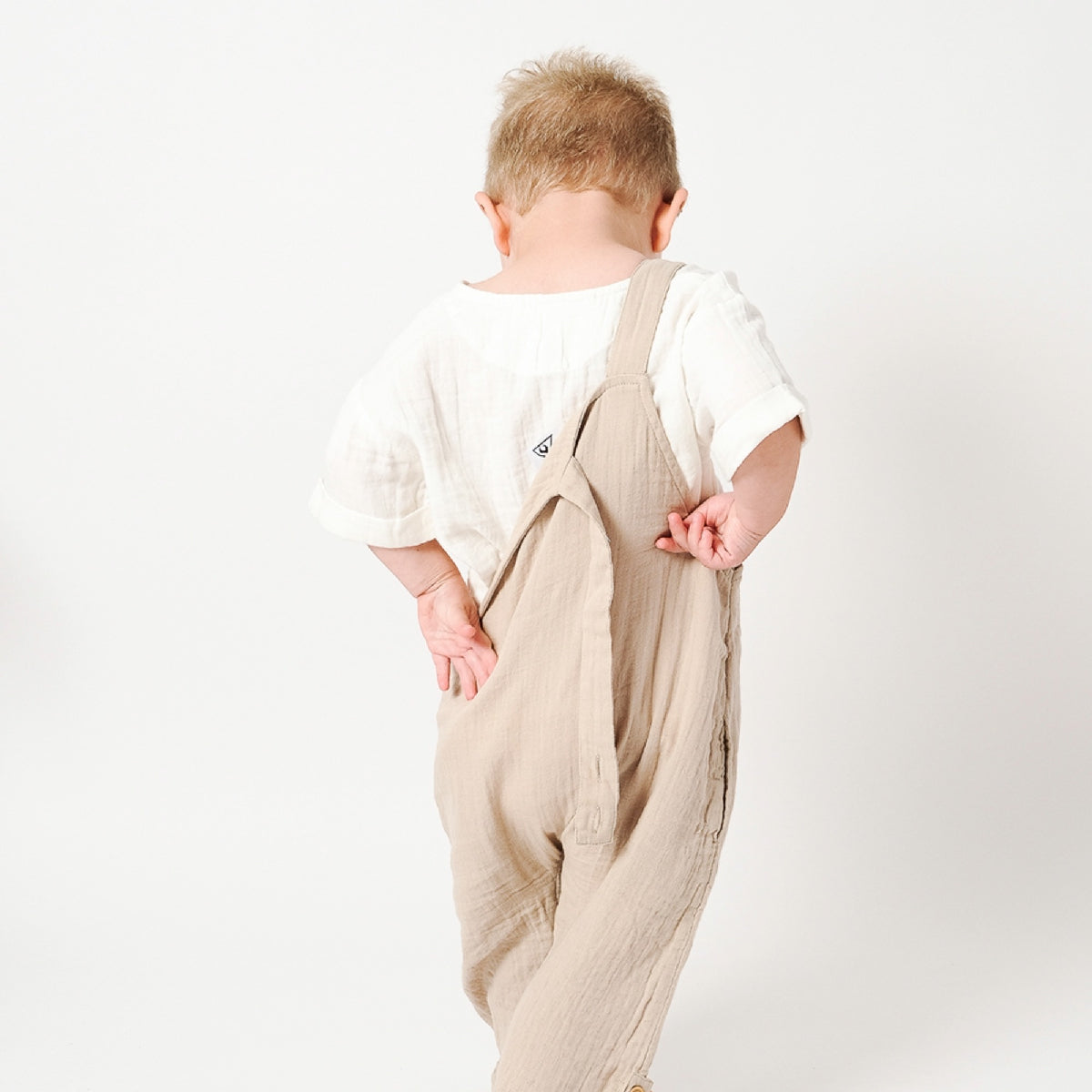 Child wearing beige overalls and a white shirt on a plain background