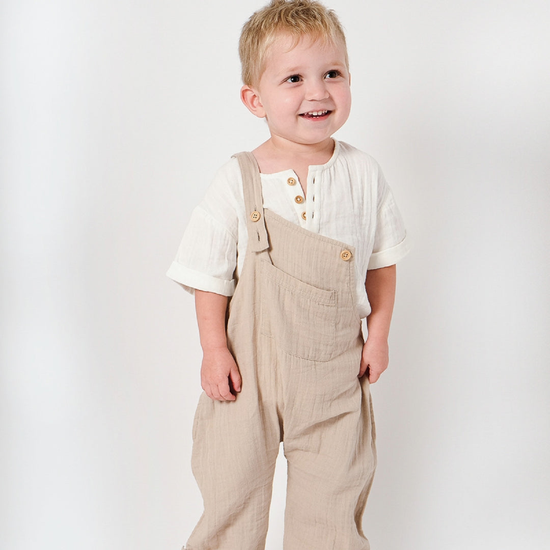 Child wearing beige overalls and a white shirt on a plain background