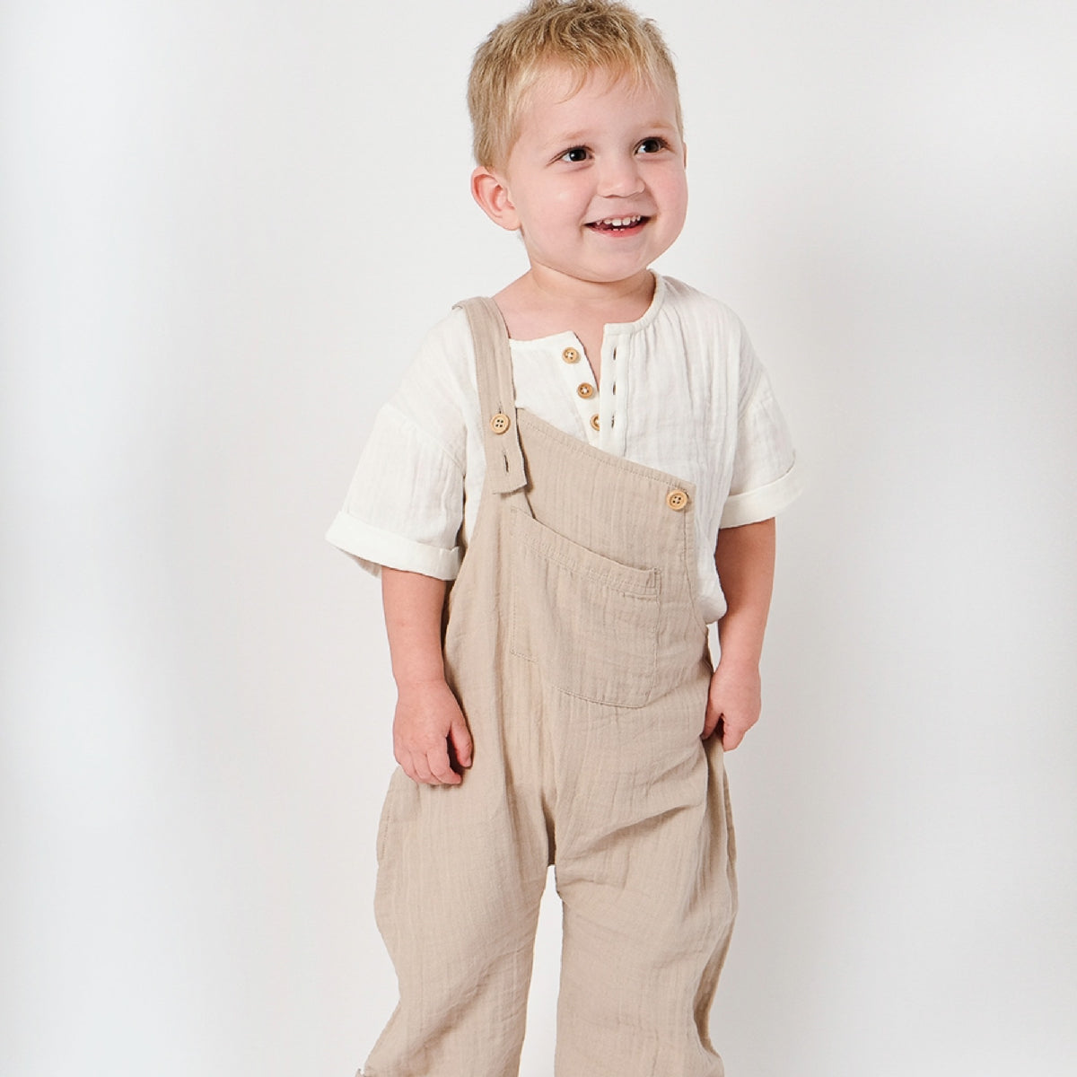 Child wearing beige overalls and a white shirt on a plain background