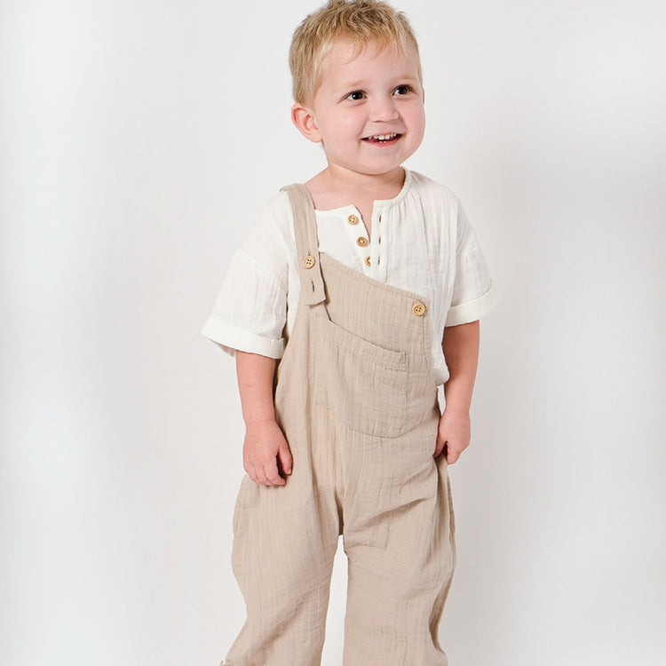 Child wearing beige overalls and a white shirt on a plain background