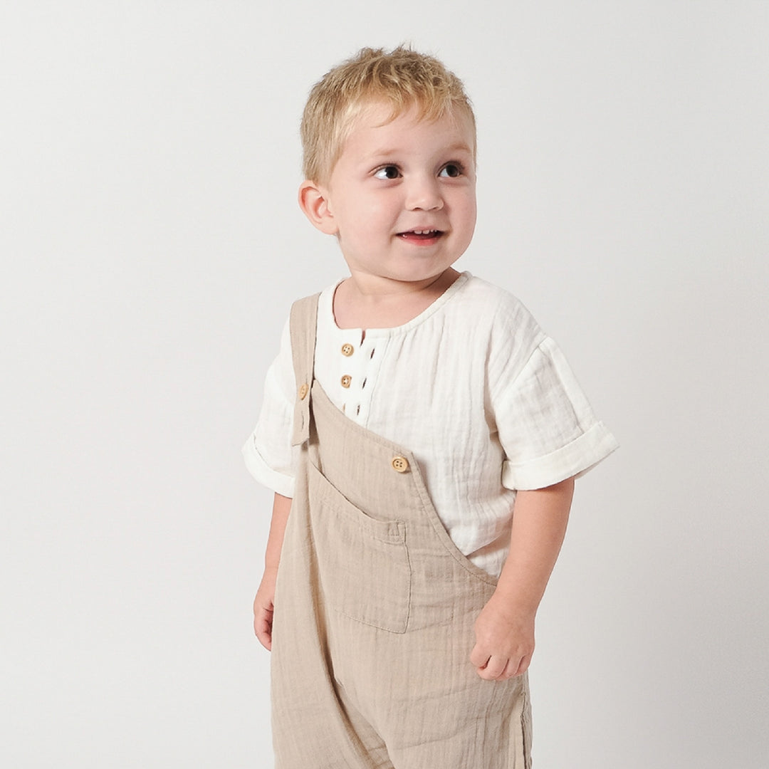 Child wearing a white shirt and beige overalls on a plain background