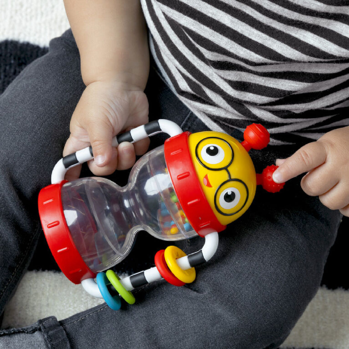 Child holding a colorful toy with a face design, sitting on a striped fabric surface.