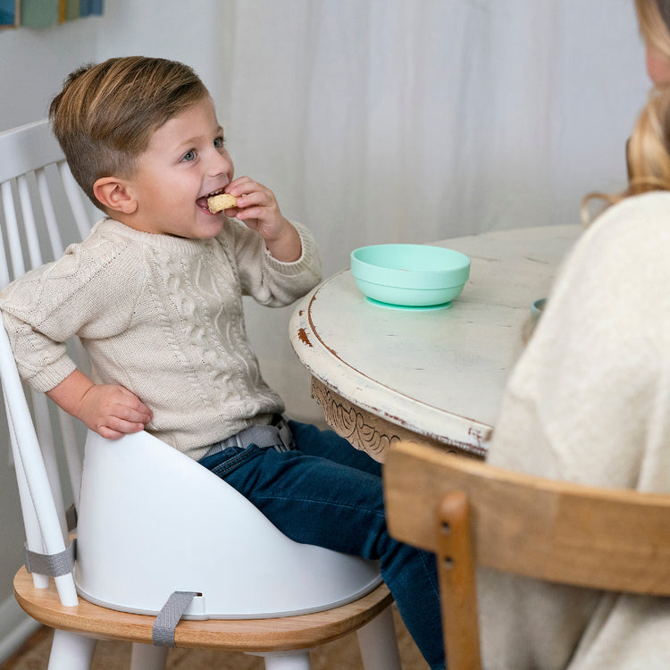 Child sitting on a chair eating with an adult at a table with a green bowl.