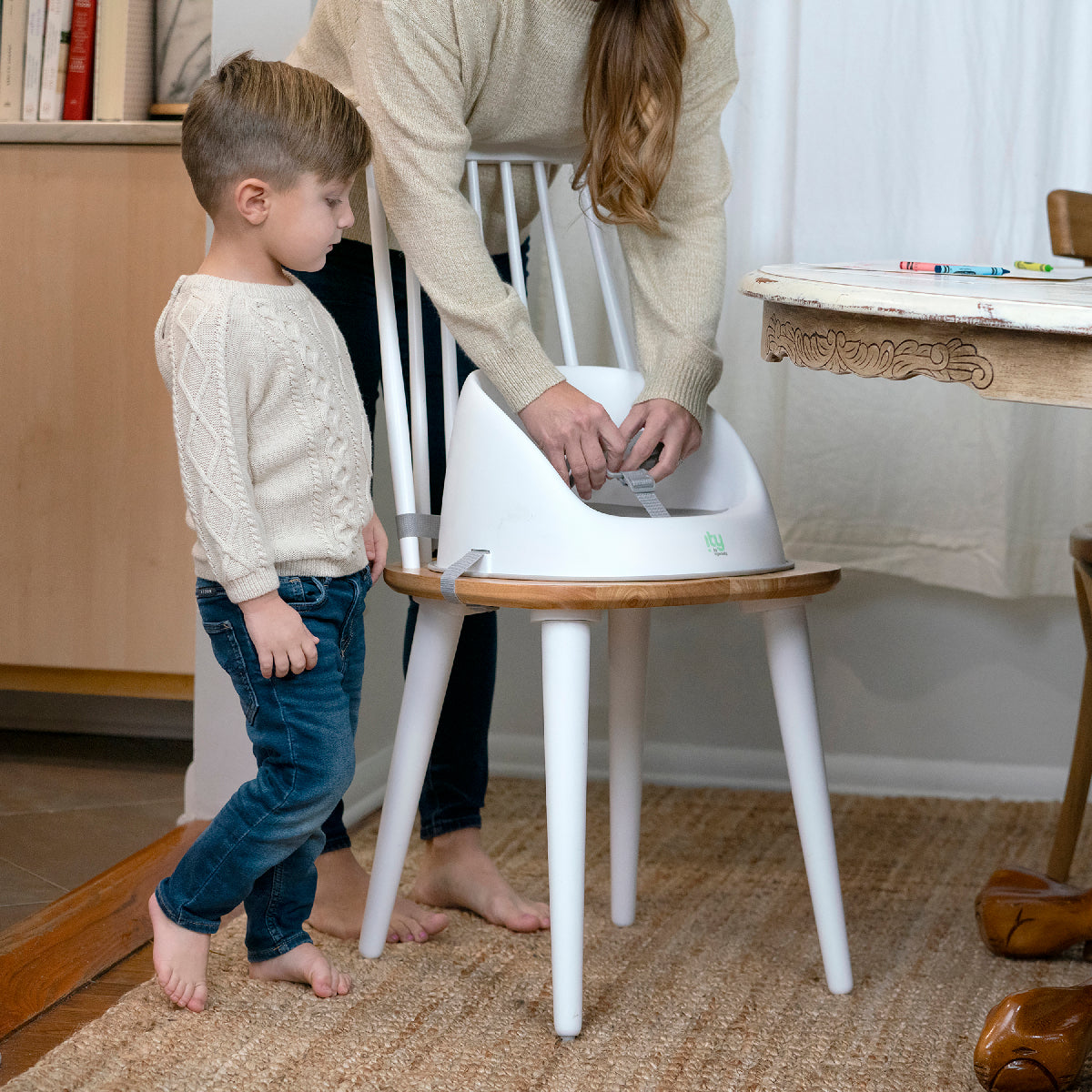 Child standing next to a chair with a white cushion, watched by an adult.