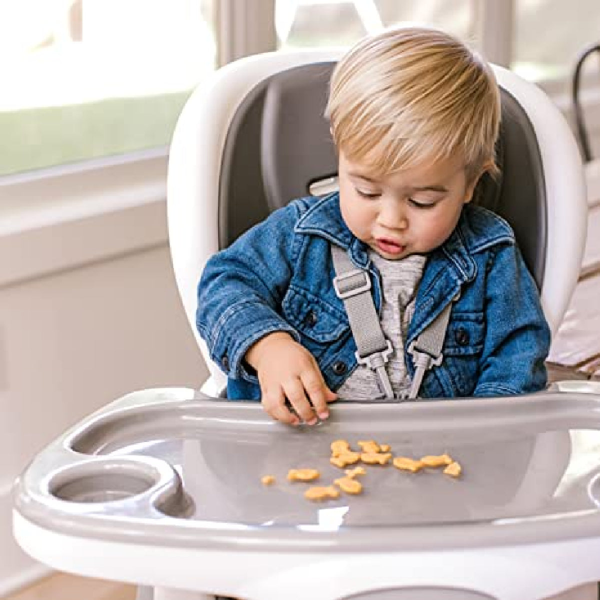 Child in a high chair with a tray, eating snacks indoors.